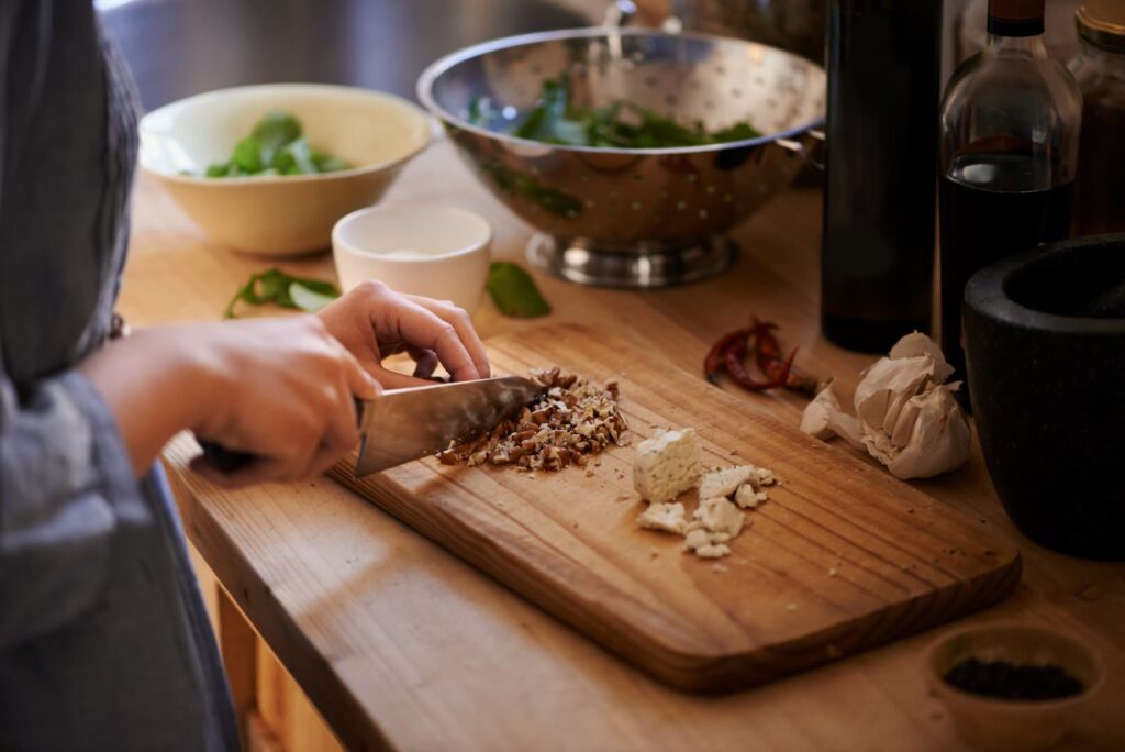 A close-up, high-angle shot of a person's hands using a chef's knife to finely chop nuts on a wooden cutting board. The rustic kitchen setting includes fresh garlic, bowls of leafy greens, and various cooking oils, emphasizing a focus on healthy, whole-food preparation.
